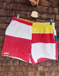 Vorderansicht der Upcycling-Frotteeshorts „Mosaic sea breeze '81“ von Old Wave auf einem Kleiderbügel vor einer Holzwand. Das asymmetrische Design zeigt ein linkes Hosenbein in Weiß und kräftigem Rot mit dem eingenähten Old Wave-Logo am Saum, kombiniert mit einem rechten Hosenbein in Gelb, Rot und Weiß.