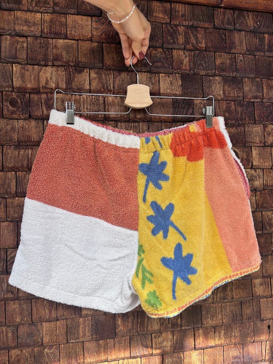 Rückansicht der Upcycling-Frotteeshorts „Mosaic stripe-wave mix '84“ von Old Wave auf einem Kleiderbügel vor einer rustikalen Holzwand. Das asymmetrische Design zeigt ein linkes Hosenbein in hellem Rotbraun und Weiß, kombiniert mit einem rechten Hosenbein in kräftigem Gelb, das mit blauen und orangefarbenen Retro-Blumenmustern verziert ist.