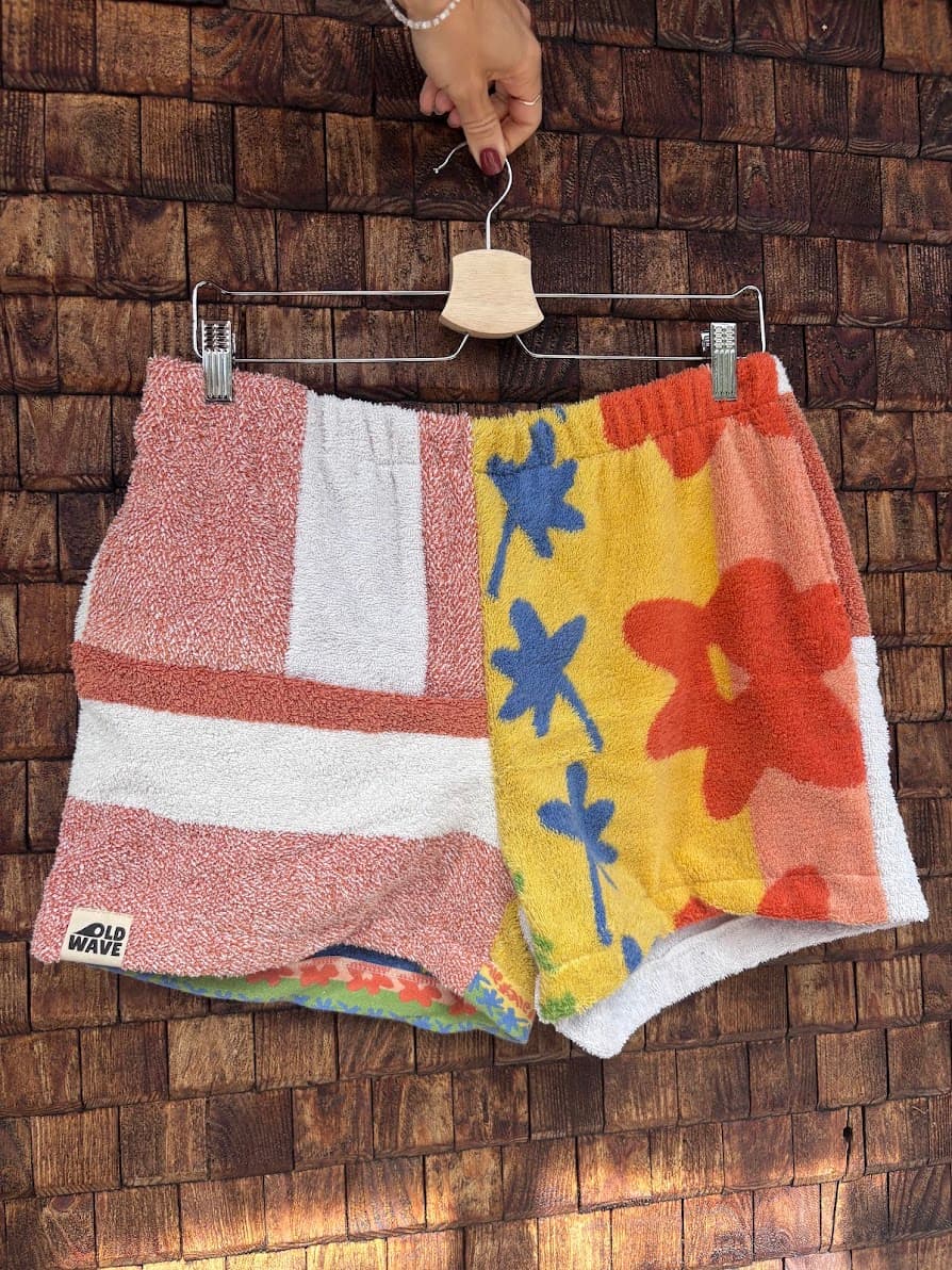 Vorderansicht der Upcycling-Frotteeshorts „Mosaic stripe-wave mix '84“ von Old Wave auf einem Kleiderbügel vor einer Holzwand. Das Patchwork-Muster zeigt auf der linken Seite rotbraune und weiße Blöcke mit dem eingenähten Old Wave-Logo am Saum, während das rechte Hosenbein das gelbe Vintage-Handtuchmaterial mit markanten blauen und orangefarbenen Blumen präsentiert.