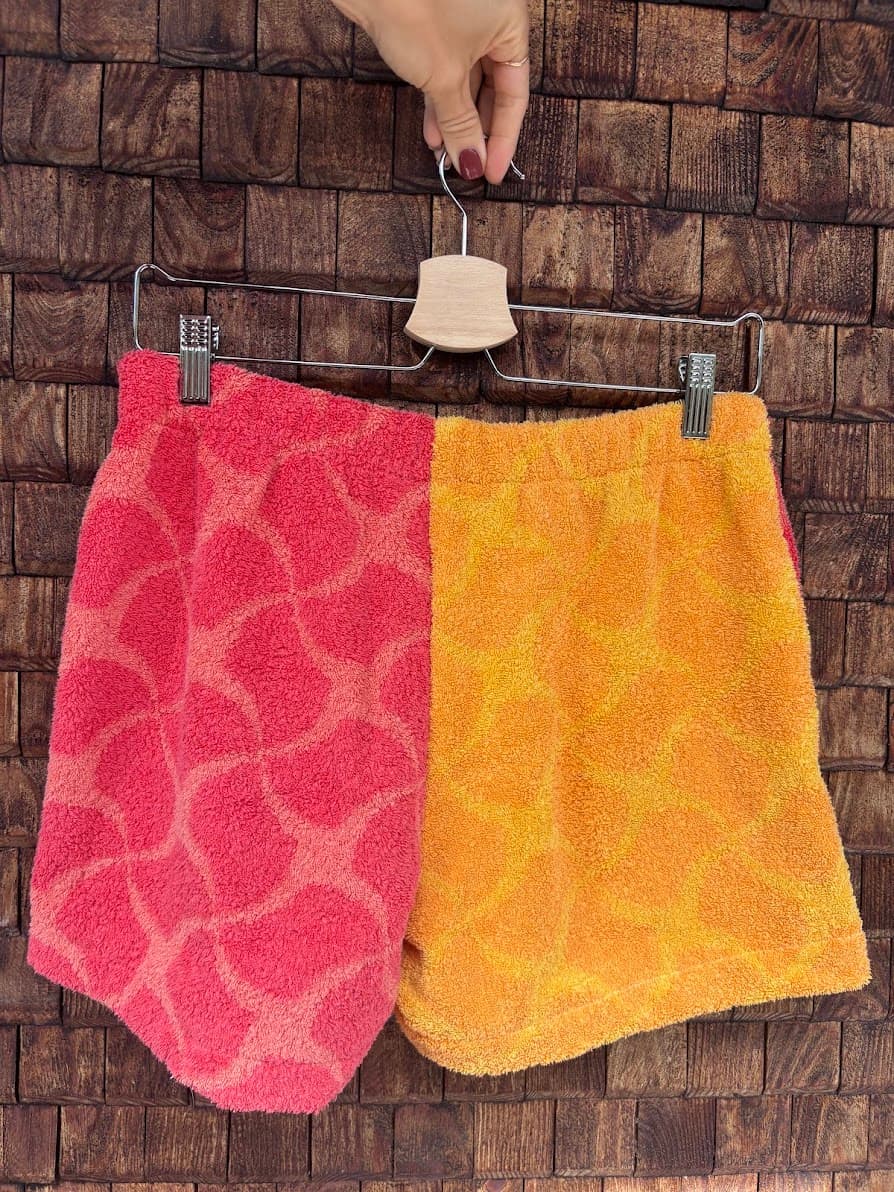 Rückansicht der Upcycling-Frotteeshorts „Mosaic sunset '79“ von Old Wave auf einem Kleiderbügel vor einer rustikalen Holzwand. Das sommerliche Design zeigt ein linkes Hosenbein in kräftigem Pink-Rot mit einem helleren Mosaikmuster und ein rechtes Hosenbein in warmem Orange mit gelbem Mosaikmuster.