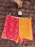 Vorderansicht der Upcycling-Frotteeshorts „Mosaic sunset '79“ von Old Wave auf einem Kleiderbügel vor einer Holzwand. Das asymmetrische Patchwork-Design spiegelt die Rückseite: Links ist das pink-rote Mosaikmuster mit dem eingenähten weißen Old Wave-Logo am Saum zu sehen, rechts das orange-gelbe Mosaikmuster.