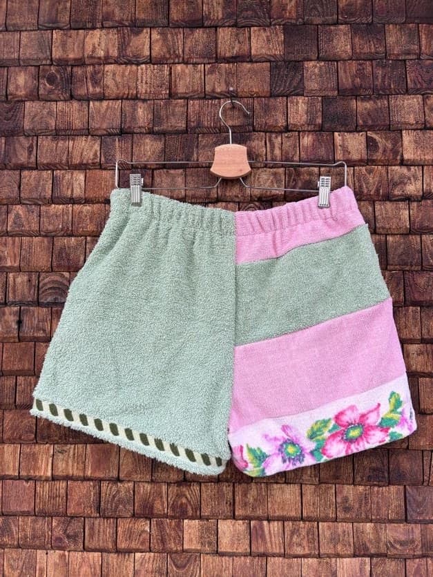 Rückansicht der Upcycling-Frotteeshorts „Moss Blossom ‘88“ von Old Wave auf einem Kleiderbügel vor einer Holzwand. Die Rückseite präsentiert ein harmonisches Patchwork-Muster mit einer unifarbenen linken Gesäßhälfte in Salbeigrün und einer rechten Gesäßhälfte, die aus breiten horizontalen Farbblöcken in Grün und Rosa besteht und am Saum mit der floralen Retro-Blumenborte abschließt.