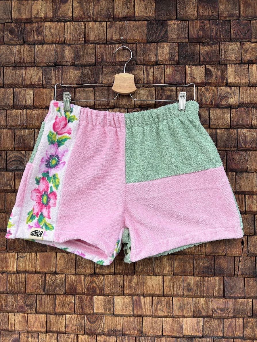 Vorderansicht der Upcycling-Frotteeshorts „Moss Blossom ‘88“ von Old Wave auf einem Kleiderbügel vor einer rustikalen Holzwand. Das verspielte Retro-Design zeigt ein rechtes Hosenbein aus pastellrosa Frottee, das an der Außenseite durch eine auffällige weiße Borte mit bunten, eingewebten Blumenmotiven in Pink, Grün und Lila verziert ist. Das linke Hosenbein ist im oberen Bereich in Salbeigrün gehalten und im unteren Bereich in Rosa abgesetzt.