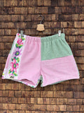 Vorderansicht der Upcycling-Frotteeshorts „Moss Blossom ‘88“ von Old Wave auf einem Kleiderbügel vor einer rustikalen Holzwand. Das verspielte Retro-Design zeigt ein rechtes Hosenbein aus pastellrosa Frottee, das an der Außenseite durch eine auffällige weiße Borte mit bunten, eingewebten Blumenmotiven in Pink, Grün und Lila verziert ist. Das linke Hosenbein ist im oberen Bereich in Salbeigrün gehalten und im unteren Bereich in Rosa abgesetzt.
