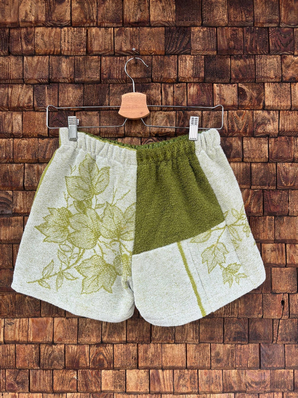 Rückansicht der Upcycling-Frotteeshorts „Moss Garden ‘84“ von Old Wave auf einem Kleiderbügel vor einer Holzwand. Die Rückseite zeigt eine asymmetrische Aufteilung: Die linke Gesäßhälfte präsentiert ein großflächiges, hellgrün gezeichnetes Vintage-Blumenmuster auf weißem Frottee. Die rechte Hälfte ist dreigeteilt – oben unifarben olivgrün, unten rechts weiß-floral gemustert und unten links mit einem schlichten weißen Frottee-Patch versehen.