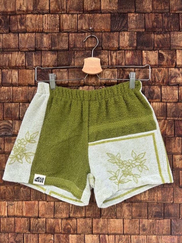 Vorderansicht der Upcycling-Frotteeshorts „Moss Garden ‘84“ von Old Wave auf einem Kleiderbügel vor einer rustikalen Holzwand. Das botanisch inspirierte Patchwork-Design besticht durch ein linkes Hosenbein aus olivgrünem Frottee mit dem Logo-Patch am Saum, abgesetzt von einem hellbeigen Seitenstreifen mit floralem Muster. Das rechte Hosenbein kombiniert ein olivgrünes Frottee-Zentrum mit einer unteren Hälfte, die ein umrahmtes, hellgrünes Blumen- und Blattmotiv auf weißem Grund zeigt.