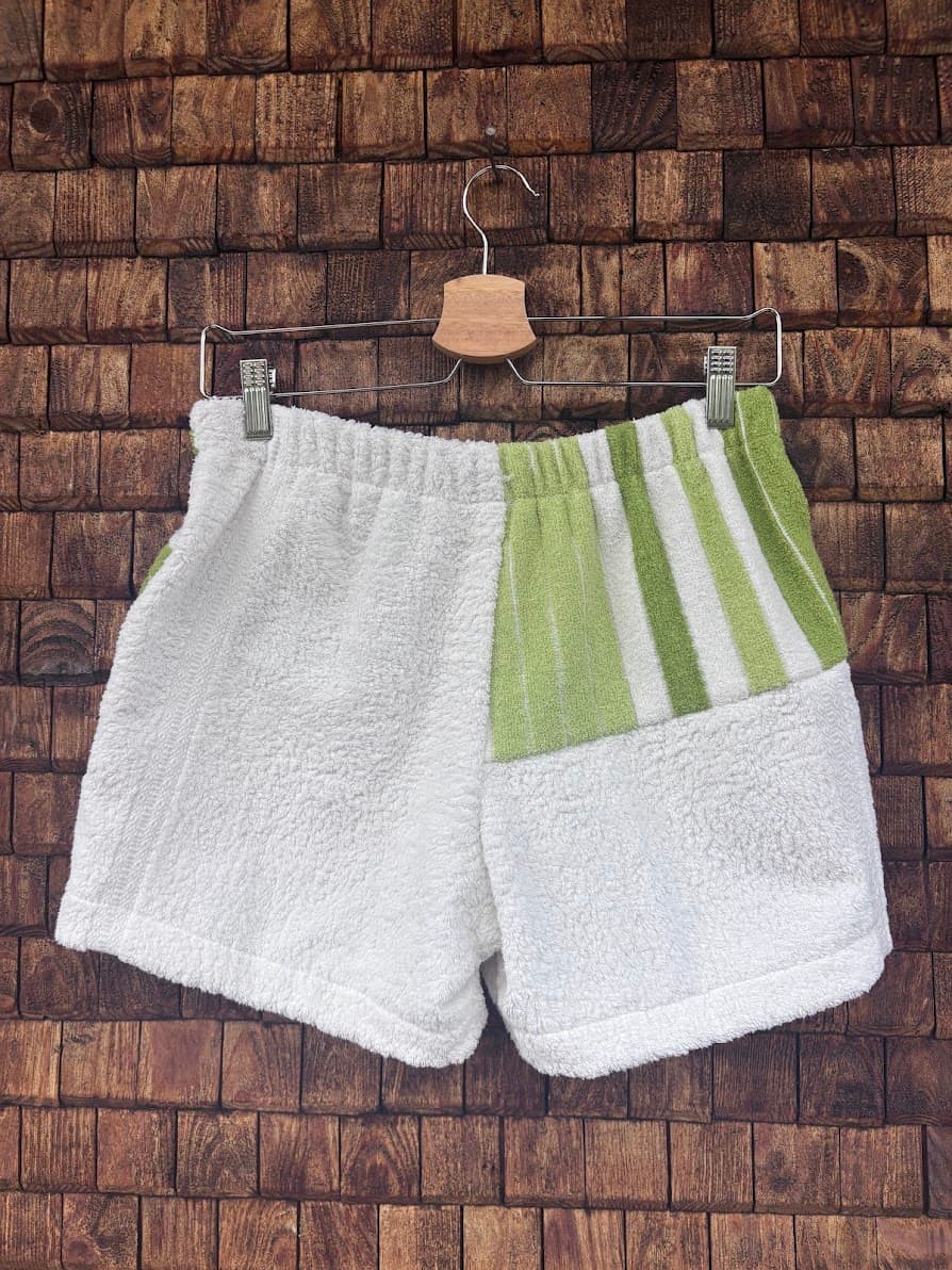 Rückansicht der Upcycling-Frotteeshorts „Moss Line ‘106“ von Old Wave auf einem Kleiderbügel vor einer Holzwand. Das Patchwork-Design präsentiert eine komplett weiße linke Gesäßhälfte, während die rechte Seite im oberen Bereich moosgrün-weiße Längsstreifen zeigt und im unteren Bereich aus weißem Vintage-Frottee besteht.