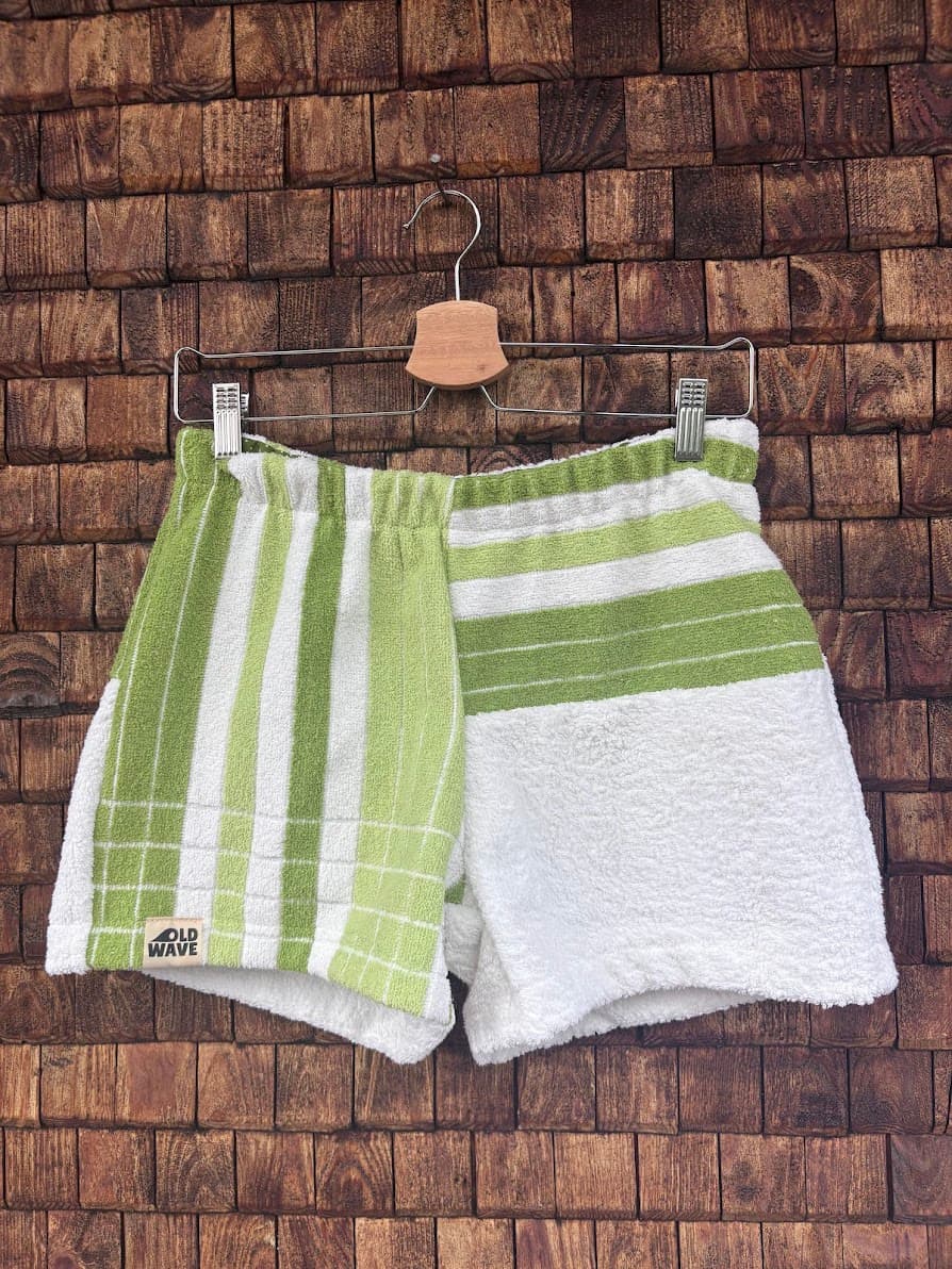 Vorderansicht der Upcycling-Frotteeshorts „Moss Line ‘106“ von Old Wave auf einem Kleiderbügel vor einer rustikalen Holzwand. Das asymmetrische Design zeigt ein linkes Hosenbein mit markanten Längsstreifen in Moosgrün und Weiß sowie dem eingenähten Old Wave-Logo am Saum, kombiniert mit einem rechten Hosenbein, das am Bund grüne Querstreifen aufweist und darunter in schlichtem Weiß gehalten ist.