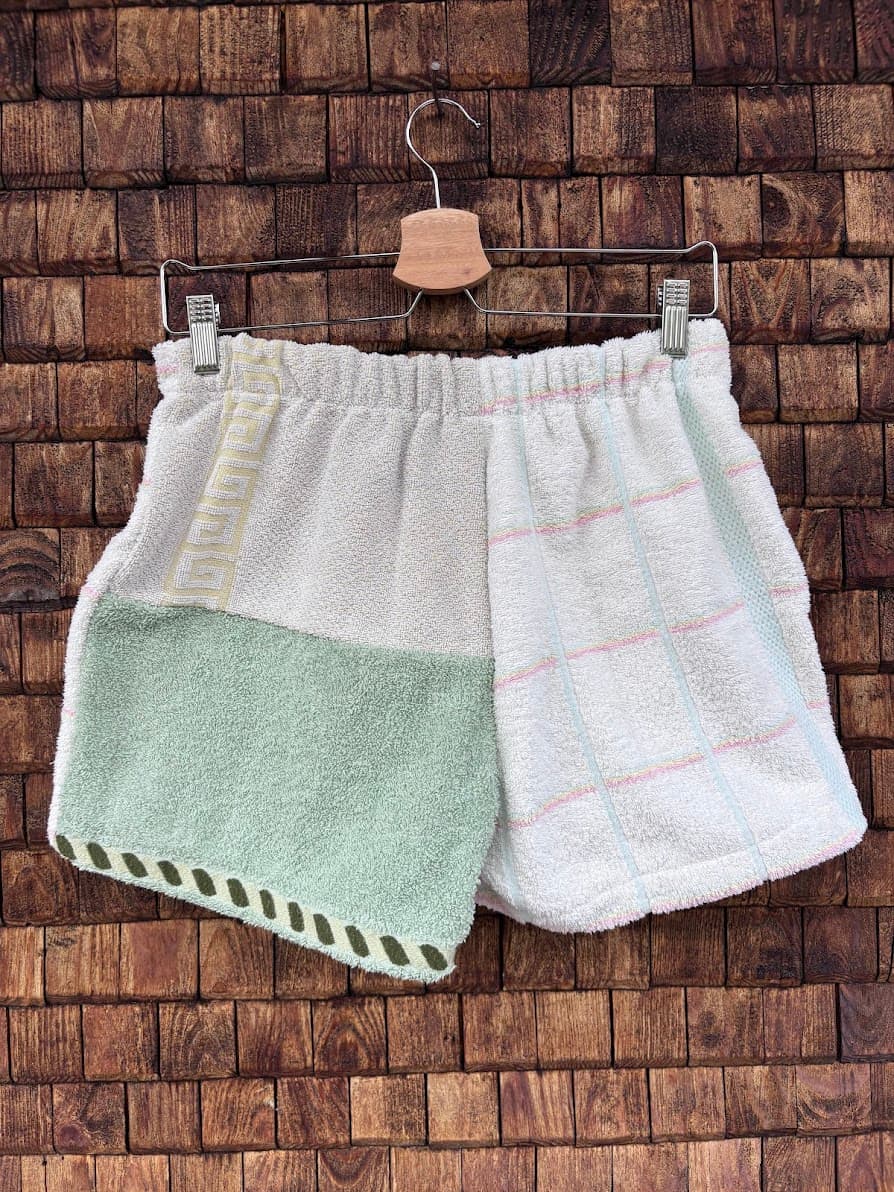 Rückansicht der Upcycling-Frotteeshorts „Moss Riviera ‘94“ von Old Wave auf einem Kleiderbügel vor einer Holzwand. Die Rückseite setzt auf ein asymmetrisches Patchwork-Muster: Das rechte Hosenbein führt das dezente, pastellfarbene Karolinienmuster fort, während die linke Gesäßhälfte zweigeteilt ist – oben in schlichtem Graustoff mit gelbem Bortenakzent am Bund und unten abgesetzt in einem sanften Moosgrün mit gemustertem Saum.
