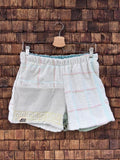 Vorderansicht der Upcycling-Frotteeshorts „Moss Riviera ‘94“ von Old Wave auf einem Kleiderbügel vor einer rustikalen Holzwand. Das helle Design präsentiert ein linkes Hosenbein aus weißem Vintage-Frottee, das im unteren Bereich mit einer gelben Mäander-Borte sowie dem eingenähten Old Wave-Logo verziert ist. Das rechte Hosenbein besticht durch ein dezentes, großkariertes Linienmuster in Pastelltönen auf weißem Grund.
