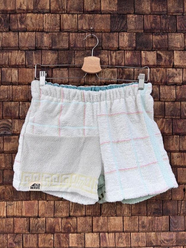 Vorderansicht der Upcycling-Frotteeshorts „Moss Riviera ‘94“ von Old Wave auf einem Kleiderbügel vor einer rustikalen Holzwand. Das helle Design präsentiert ein linkes Hosenbein aus weißem Vintage-Frottee, das im unteren Bereich mit einer gelben Mäander-Borte sowie dem eingenähten Old Wave-Logo verziert ist. Das rechte Hosenbein besticht durch ein dezentes, großkariertes Linienmuster in Pastelltönen auf weißem Grund.