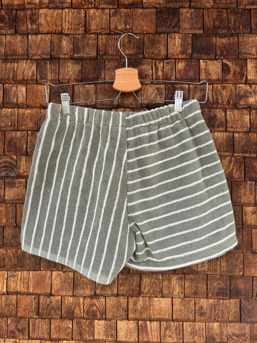 Rückansicht der Upcycling-Frotteeshorts „Olive Wave ‘74“ von Old Wave auf einem Kleiderbügel vor einer Holzwand. Die Rückseite besticht durch ein raffiniertes, geometrisches Streifenspiel in Olivgrün und Weiß: Die linke Gesäßhälfte ist durchgehend vertikal gestreift, während die rechte Hälfte ein horizontales Streifenmuster zeigt.