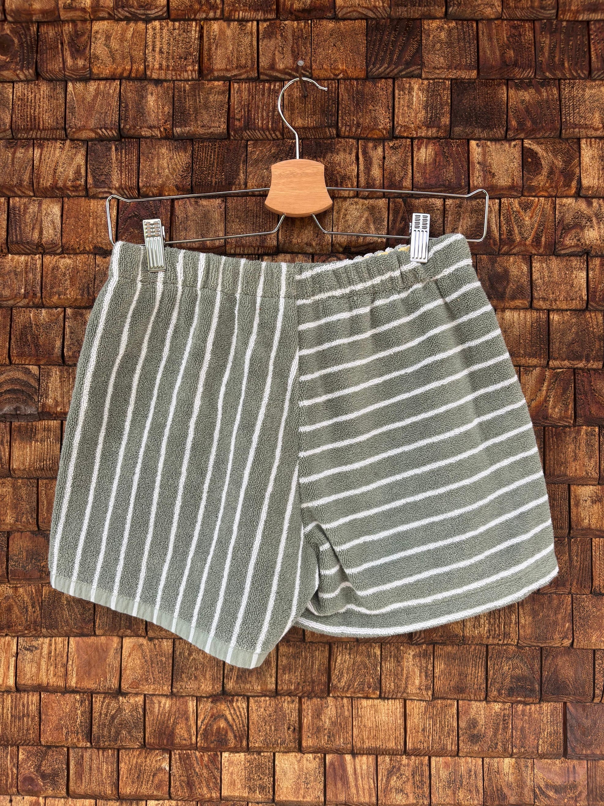 Rückansicht der Upcycling-Frotteeshorts „Olive Wave ‘74“ von Old Wave auf einem Kleiderbügel vor einer Holzwand. Die Rückseite besticht durch ein raffiniertes, geometrisches Streifenspiel in Olivgrün und Weiß: Die linke Gesäßhälfte ist durchgehend vertikal gestreift, während die rechte Hälfte ein horizontales Streifenmuster zeigt.