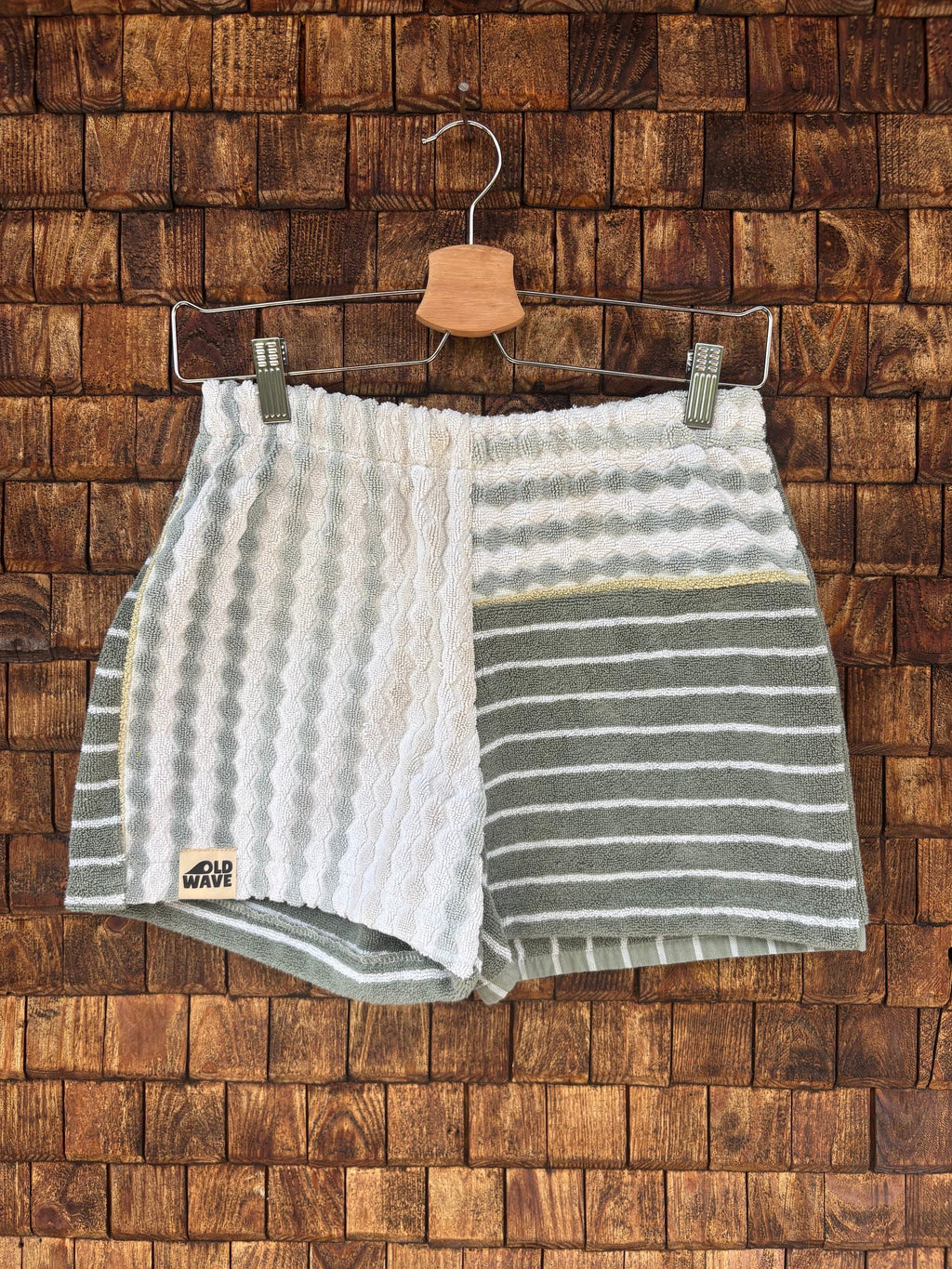 Vorderansicht der Upcycling-Frotteeshorts „Olive Wave ‘74“ von Old Wave auf einem Kleiderbügel vor einer rustikalen Holzwand. Das Patchwork-Design präsentiert ein linkes Hosenbein aus weißem Vintage-Frottee mit einer markanten, vertikalen Wellen-Reliefstruktur und dem Old Wave-Logo am Saum. Das rechte Hosenbein kombiniert einen hell gestreiften Bundbereich mit einem olivgrünen Hauptteil, der mit feinen, horizontalen weißen Streifen versehen ist.