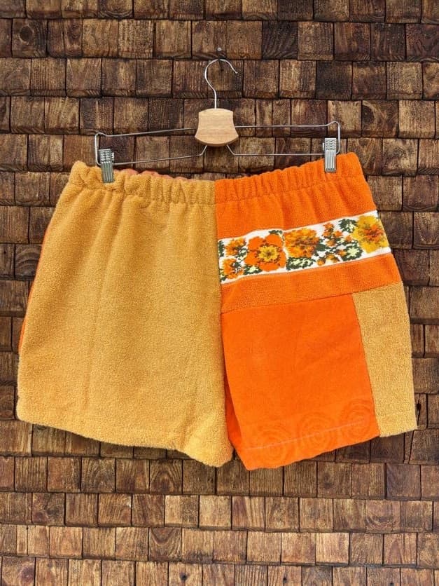 Rückansicht der Upcycling-Frotteeshorts „Orange Border ‘65“ von Old Wave auf einem Kleiderbügel vor einer Holzwand. Die Rückseite präsentiert ein harmonisches, hälftig geteiltes Color-Blocking: Die linke Gesäßhälfte besteht komplett aus senfgelbem Frotteestoff, während die rechte Hälfte in kräftigem Orange gehalten ist und im oberen Drittel durch den horizontal verlaufenden Streifen der floralen Vintage-Blumenborte akzentuiert wird.