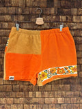 Vorderansicht der Upcycling-Frotteeshorts „Orange Border ‘65“ von Old Wave auf einem Kleiderbügel vor einer rustikalen Holzwand. Das warme Retro-Design im 60er-Jahre-Look zeigt ein linkes Hosenbein aus leuchtend orangefarbenem Frottee, das am Saum mit einer floralen Borte (Border) in Orange- und Gelbtönen sowie dem Logo-Patch verziert ist. Das rechte Hosenbein kombiniert eine obere Hälfte in Senfgelb mit einer unteren Hälfte in Orange.