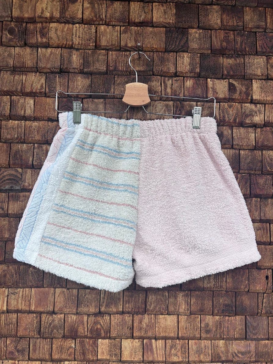 Rückansicht der Upcycling-Frotteeshorts „Pastel Breeze ‘98“ von Old Wave auf einem Kleiderbügel vor einer Holzwand. Die Rückseite spiegelt das harmonische Retro-Design wider: Die linke Gesäßhälfte zeigt die feine, pastellfarbene Streifenstruktur auf weißem Grund mit einem hellblauen Seitenstreifen, während das rechte Hosenbein schlicht aus unifarbenem, pastellrosa Frotteestoff gefertigt ist.