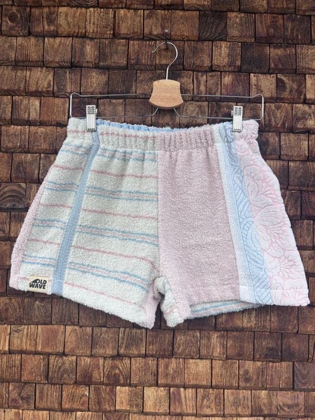 Vorderansicht der Upcycling-Frotteeshorts „Pastel Breeze ‘98“ von Old Wave auf einem Kleiderbügel vor einer rustikalen Holzwand. Das asymmetrische Patchwork-Design kombiniert ein linkes Hosenbein aus weißem Vintage-Frottee mit feinen hellblauen und lachsfarbenen Querstreifen sowie dem eingenähten Old Wave-Logo am Saum, während das rechte Hosenbein in sanftem Pastellrosa gehalten ist und einen vertikalen hellblauen Streifenakzent am Rand besitzt.