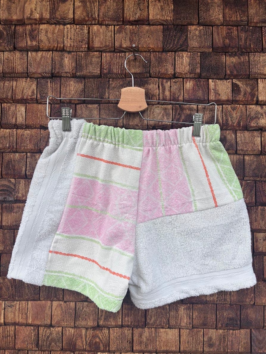 Rückansicht der Upcycling-Frotteeshorts „Pastel Diamond ‘104“ von Old Wave auf einem Kleiderbügel vor einer Holzwand. Das asymmetrische Design zeigt ein linkes Hosenbein mit hellgrünen und rosa Streifenmustern auf weißem Grund, während die rechte Gesäßhälfte vorwiegend aus schlichtem, weißem Frotteestoff gefertigt ist.