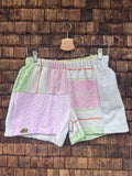 Vorderansicht der Upcycling-Frotteeshorts „Pastel Diamond ‘104“ von Old Wave auf einem Kleiderbügel vor einer rustikalen Holzwand. Das zarte Patchwork-Design zeigt ein linkes Hosenbein in hellem Flieder-Rosa mit dem eingenähten Old Wave-Logo am Saum, kombiniert mit einem rechten Hosenbein aus weißem Vintage-Frottee, das durch pastellgrüne, rosa und orangefarbene Streifenakzente ergänzt wird.