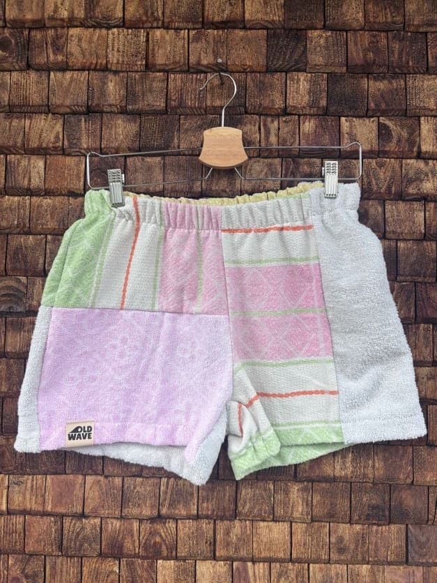 Vorderansicht der Upcycling-Frotteeshorts „Pastel Diamond ‘104“ von Old Wave auf einem Kleiderbügel vor einer rustikalen Holzwand. Das zarte Patchwork-Design zeigt ein linkes Hosenbein in hellem Flieder-Rosa mit dem eingenähten Old Wave-Logo am Saum, kombiniert mit einem rechten Hosenbein aus weißem Vintage-Frottee, das durch pastellgrüne, rosa und orangefarbene Streifenakzente ergänzt wird.