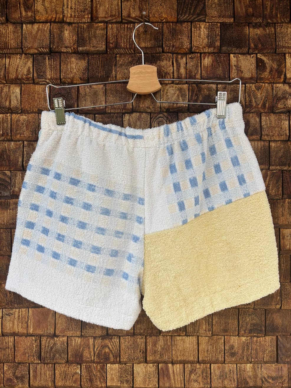 Rückansicht der Upcycling-Frotteeshorts „Pastel Paisley ‘79“ von Old Wave auf einem Kleiderbügel vor einer Holzwand. Die Rückseite überrascht mit einem kontrastierenden Patchwork-Muster: Die linke Gesäßseite zeigt ein blau-weißes Karomuster auf weißem Frottee. Die rechte Seite ist zweigeteilt – oben mit dem gleichen blau-weißen Karomuster und unten abgesetzt in einem unifarbenen, pastellgelben Frotteestoff.