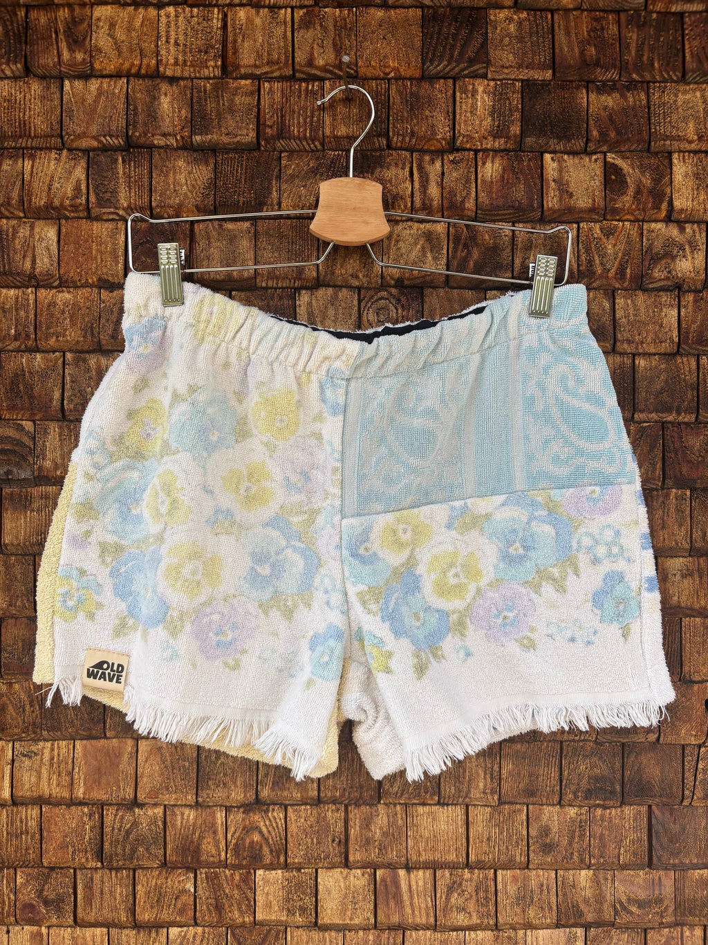 Vorderansicht der Upcycling-Frotteeshorts „Pastel Paisley ‘79“ von Old Wave auf einem Kleiderbügel vor einer rustikalen Holzwand. Das verspielte Retro-Design im pastelligen Look zeigt ein linkes Hosenbein mit einem gelb-blauen Blumenmuster auf weißem Grund und das Old Wave-Logo am gefransten Saum. Das rechte Hosenbein kombiniert im oberen Bereich ein hellblaues Paisley-Muster mit dem passenden Blumenprint im unteren Teil.