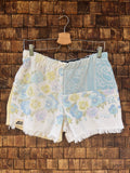 Vorderansicht der Upcycling-Frotteeshorts „Pastel Paisley ‘79“ von Old Wave auf einem Kleiderbügel vor einer rustikalen Holzwand. Das verspielte Retro-Design im pastelligen Look zeigt ein linkes Hosenbein mit einem gelb-blauen Blumenmuster auf weißem Grund und das Old Wave-Logo am gefransten Saum. Das rechte Hosenbein kombiniert im oberen Bereich ein hellblaues Paisley-Muster mit dem passenden Blumenprint im unteren Teil.