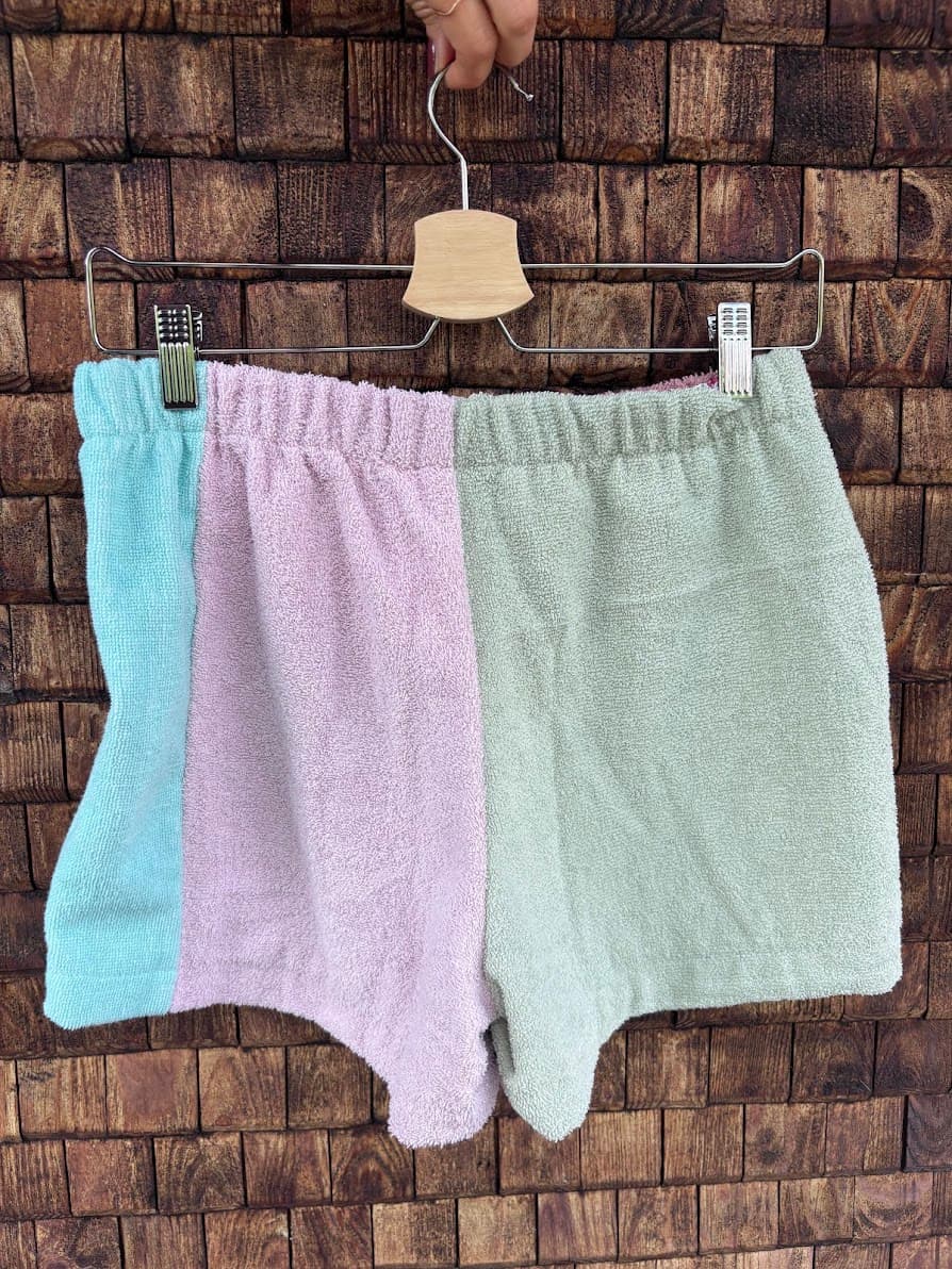 Rückansicht der Frottee-Patchwork-Shorts „Peach '70“ von Old Wave an einem Kleiderbügel. Diese Seite zeigt ein asymmetrisches Gesäßdesign aus upgecycelten Handtüchern: Das linke Hosenbein wechselt von einem mintblauen Außenstreifen zu einer altrosafarbenen Fläche, während das rechte Hosenbein komplett aus einem sanften, mintgrünen Frotteestoff gefertigt ist.
