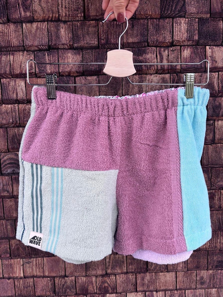 Upcycling-Shorts „Peach '70“ von Old Wave auf einem Kleiderbügel vor einer dunklen Holzwand. Das dreigeteilte Patchwork-Design aus Frottee-Handtüchern kombiniert ein rechtes Hosenbein in hellem Altrosa mit einem hellgrauen Hosenbein links, das im oberen Bereich ebenfalls altrosa gehalten ist und an einen seitlichen Streifen in Mintblau grenzt.