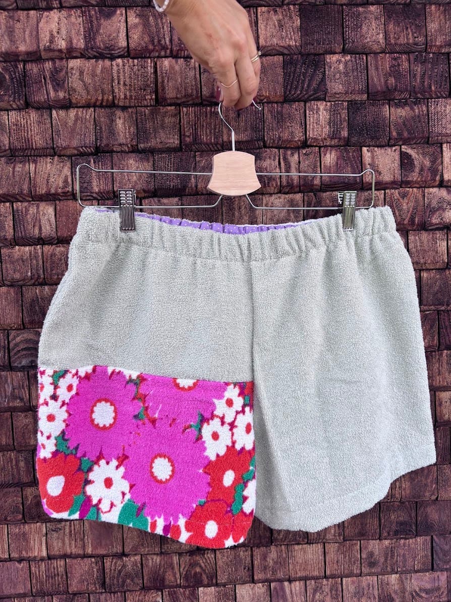 Rückansicht der Frottee-Patchwork-Shorts „Pistachio '77“ von Old Wave an einem Kleiderbügel. Diese Perspektive zeigt die umgekehrte Stoffkombination: Das linke Hosenbein besteht durchgehend aus dem sanften, hellgrau-grünen Frotteestoff, während das rechte Hosenbein im unteren Drittel mit einem auffälligen, knalligen Vintage-Blumenmuster in Pink, Rot und Weiß abschließt.