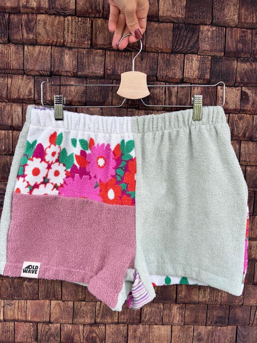 Upcycling-Shorts „Pistachio '77“ von Old Wave auf einem Kleiderbügel vor einer dunklen Holzwand. Das Patchwork-Design aus Frottee-Handtüchern kombiniert ein komplett hellgrau-grünes Hosenbein auf der rechten Seite mit einem gemusterten linken Hosenbein. Dieses zeigt im oberen Bereich ein buntes, leuchtendes Retromuster mit pinken und roten Blumen auf weißem Grund und im unteren Teil eine altrosafarbene Frotteefläche.