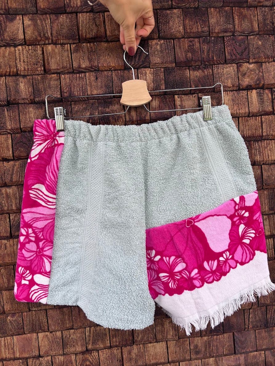 Rückansicht der Frottee-Patchwork-Shorts „Positano '75“ von Old Wave an einem Kleiderbügel. Diese Perspektive zeigt die Rückseite mit einer vorwiegend mintgrünen Gesäßpartie, die links von dem vertikalen, pinken Vintage-Blumenmuster flankiert wird, während das rechte Hosenbein im unteren Drittel mit einem auffälligen, intensiv pinken Blumen- und Rankenmotiv sowie einem weißen Fransensaum abschließt.