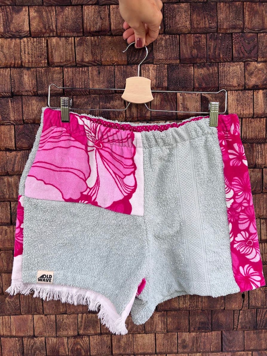 Upcycling-Shorts „Positano '75“ von Old Wave auf einem Kleiderbügel vor einer dunklen Holzwand. Das Patchwork-Design kombiniert ein komplett mintfarbenes Hosenbein auf der rechten Seite mit einem gemusterten linken Hosenbein. Dieses zeigt oben eine großflächige, weiße Vintage-Fläche mit pinken Hibiskusblüten, unten mintfarbenen Frotteestoff mit Fransen am Saum und wird an der Seite von einer leuchtend pink-floralen Stoffbahn abgeschlossen.