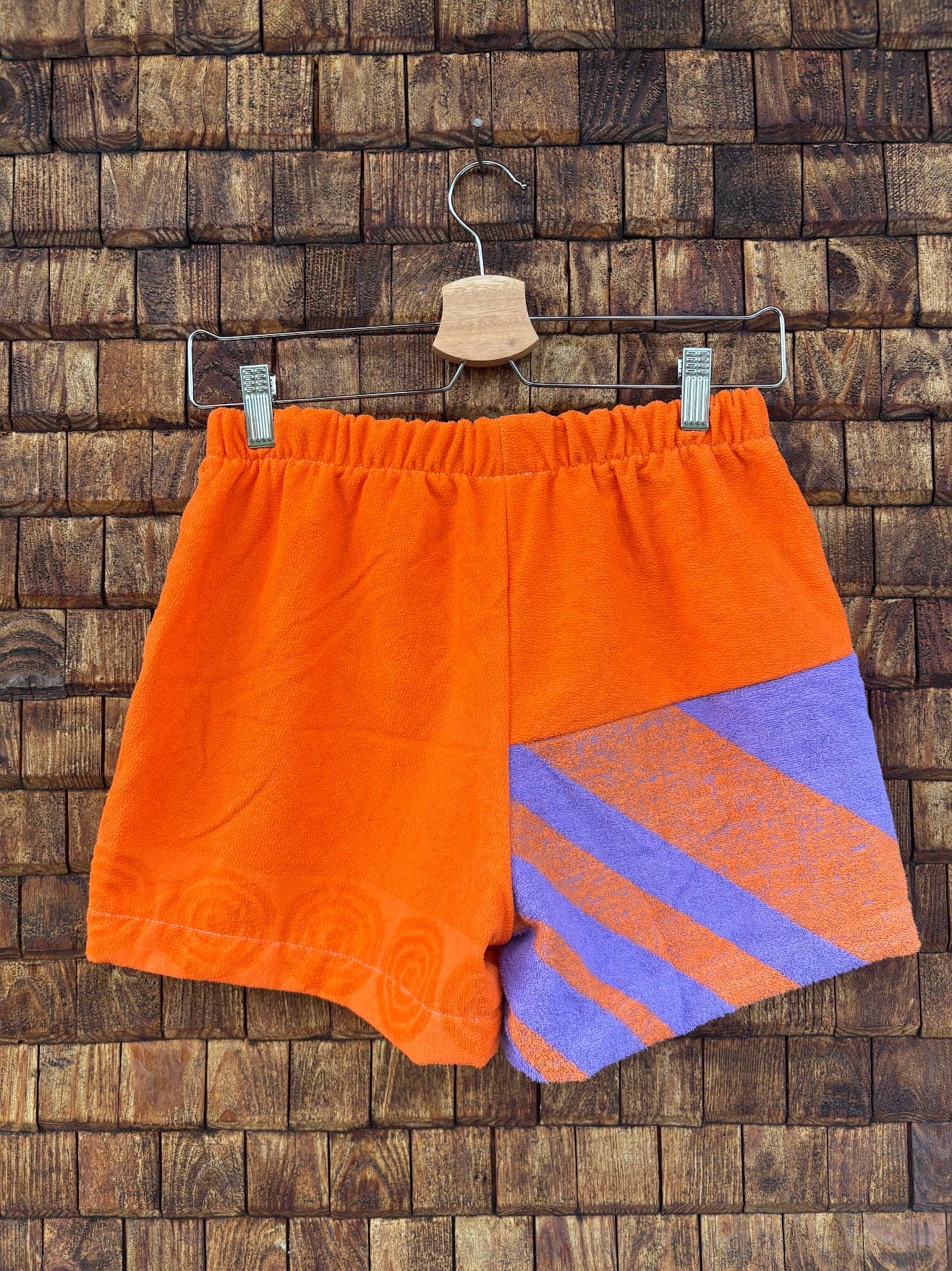Rückansicht der Upcycling-Frotteeshorts „Purple Sunset ‘83“ von Old Wave auf einem Kleiderbügel vor einer Holzwand. Die Rückseite zeigt ein klares, asymmetrisches Color-Blocking-Design: Die linke Gesäßhälfte besteht vollständig aus unifarbenem, kräftig orangefarbenem Frotteestoff, während auf der rechten Gesäßhälfte im unteren Bereich ein geometrisches Panel mit diagonalen lila-orangen Retro-Streifen aufgenäht ist.