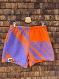 Vorderansicht der Upcycling-Frotteeshorts „Purple Sunset ‘83“ von Old Wave auf einem Kleiderbügel vor einer rustikalen Holzwand. Das sportliche Retro-Design im 80er-Jahre-Stil kombiniert leuchtendes Orange mit lilafarbenen Akzenten. Das rechte Hosenbein ist größtenteils in Lila mit orangen Streifenelementen gehalten und trägt das Logo-Patch am Saum. Das linke Hosenbein zeigt ein asymmetrisch angesetztes, lila-orange diagonal gestreiftes Musterpanel im unteren Drittel.