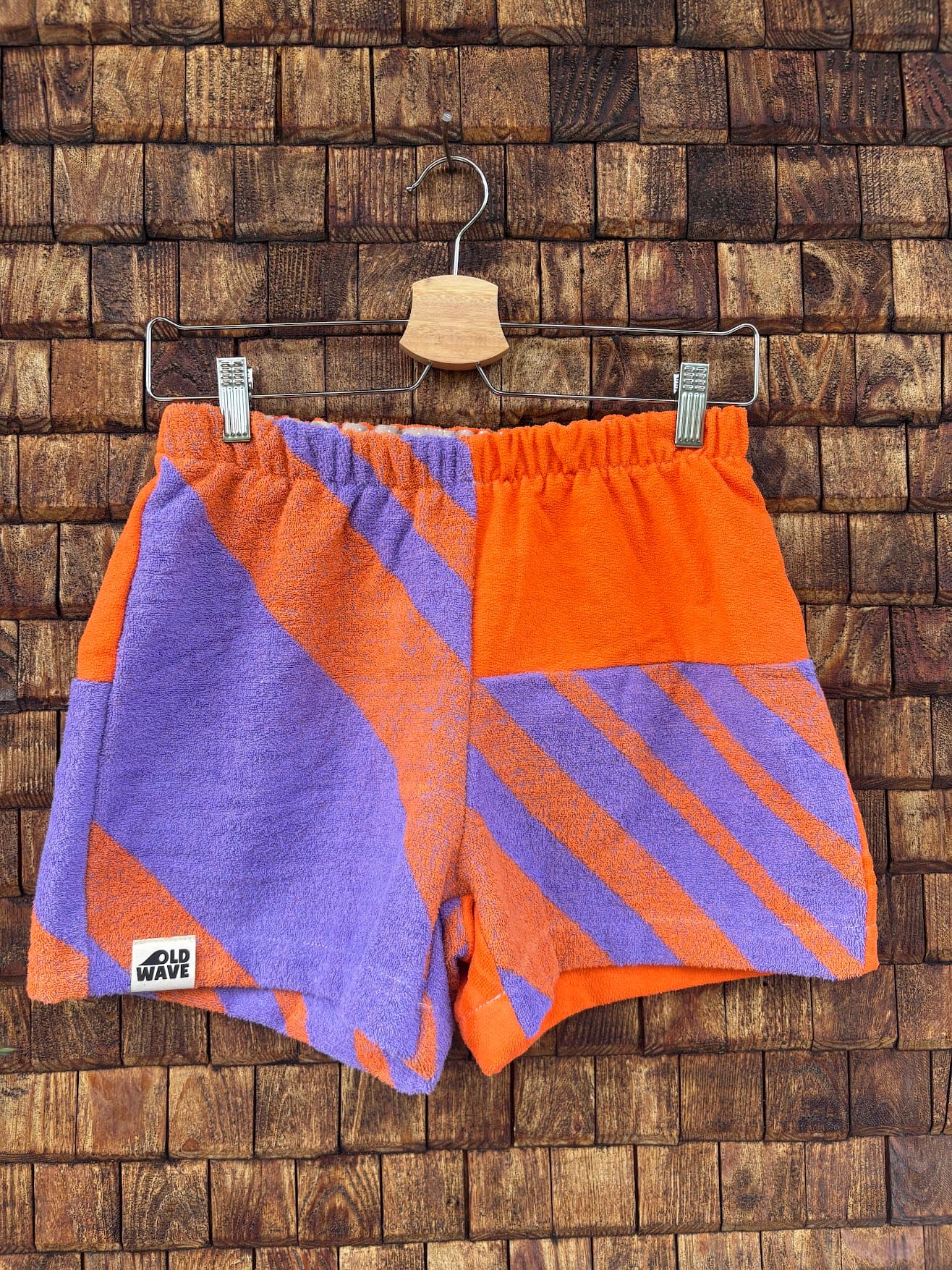 Vorderansicht der Upcycling-Frotteeshorts „Purple Sunset ‘83“ von Old Wave auf einem Kleiderbügel vor einer rustikalen Holzwand. Das sportliche Retro-Design im 80er-Jahre-Stil kombiniert leuchtendes Orange mit lilafarbenen Akzenten. Das rechte Hosenbein ist größtenteils in Lila mit orangen Streifenelementen gehalten und trägt das Logo-Patch am Saum. Das linke Hosenbein zeigt ein asymmetrisch angesetztes, lila-orange diagonal gestreiftes Musterpanel im unteren Drittel.