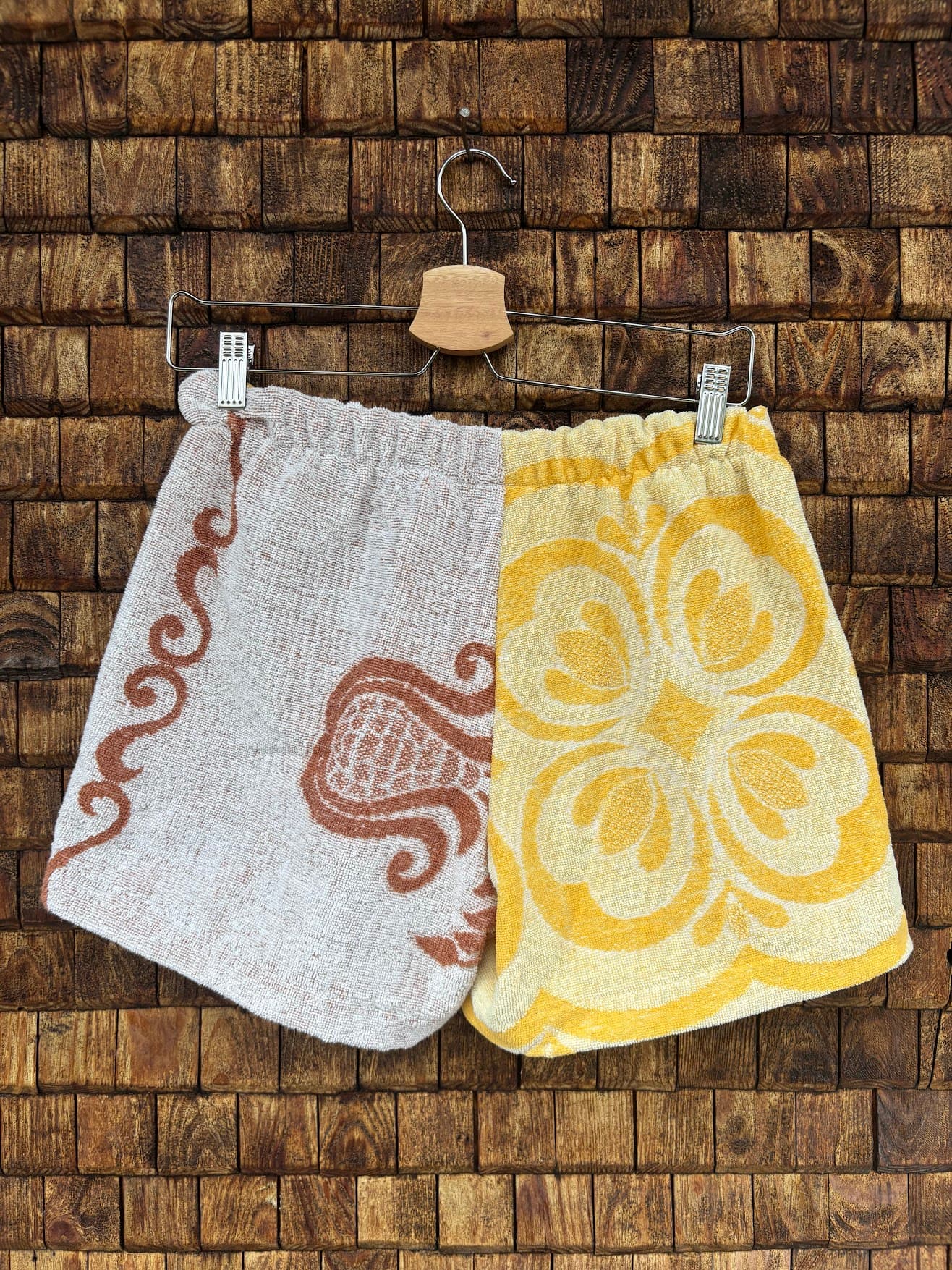 Rückansicht der Upcycling-Frotteeshorts „Retro Blossom ‘68“ von Old Wave auf einem Kleiderbügel vor einer Holzwand. Die Rückseite führt das zweigeteilte Konzept harmonisch fort. Die linke Gesäßhälfte besteht aus weißem Frotteestoff, der an der Außennaht mit einer eleganten, braun geschwungenen Ornament-Borte verziert ist, während die rechte Hälfte vollständig im strahlend gelb-goldenen Retro-Blumenmuster gehalten ist.