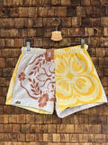 Vorderansicht der Upcycling-Frotteeshorts „Retro Blossom ‘68“ von Old Wave auf einem Kleiderbügel vor einer rustikalen Holzwand. Das charmante Vintage-Design im Look der 60er-Jahre ist hälftig geteilt: Das linke Hosenbein zeigt ein filigranes, terracottabraunes Blumen- und Ornamentmuster auf weißem Frottee sowie das Old Wave-Logo am Saum. Das rechte Hosenbein besticht durch ein großflächiges, leuchtend gelbes Retro-Blütenmuster.