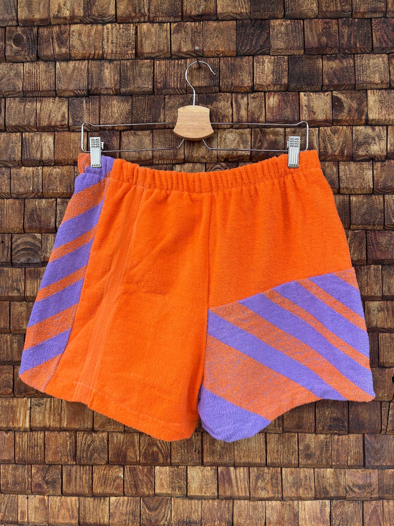 Rückansicht der Upcycling-Frotteeshorts „Retro Chevron ‘81“ von Old Wave auf einem Kleiderbügel vor einer Holzwand. Die Rückseite spiegelt das dynamische Geometric-Design wider: Die linke Gesäßhälfte ist in hellem Orange gehalten und zeigt am äußeren Rand diagonale lila Streifen. Die rechte Gesäßhälfte setzt im unteren Bereich das asymmetrisch angesetzte, lila-orangefarbene Retro-Streifenmuster fort.