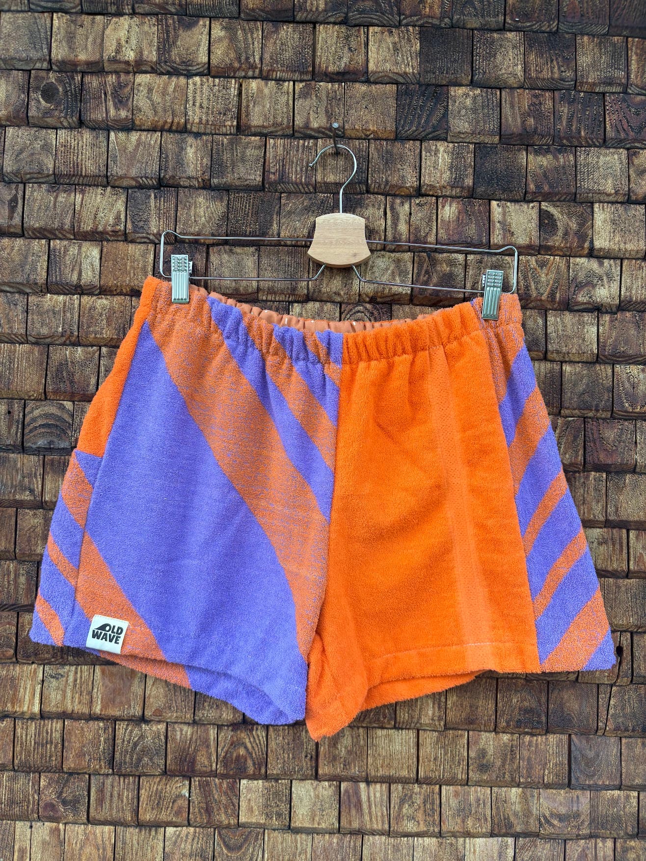 Vorderansicht der Upcycling-Frotteeshorts „Retro Chevron ‘81“ von Old Wave auf einem Kleiderbügel vor einer rustikalen Holzwand. Das sportliche Retro-Design kombiniert leuchtendes Orange mit einem markanten, lila-orange gestreiften Chevron-Muster im 80er-Jahre-Stil. Das rechte Hosenbein besticht durch das großflächige, diagonale Streifenmuster und trägt das Old Wave-Logo am Saum, während das linke Hosenbein größtenteils orange ist und an der Außenseite lila Streifenakzente zeigt.