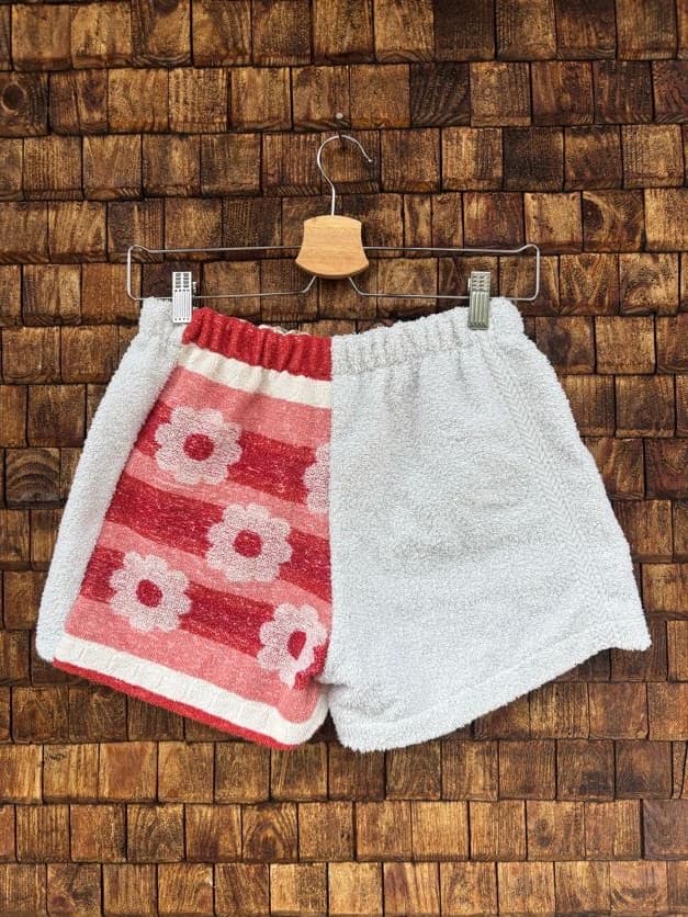Rückansicht der Upcycling-Frotteeshorts „Retro Stripes ‘77“ von Old Wave auf einem Kleiderbügel vor einer Holzwand. Die Rückseite teilt sich hälftig in eine schlichte, hellgrau-weiße Frotteeseite rechts und eine linke Gesäßhälfte, die komplett aus dem roten Vintage-Frotteestoff mit weißen Retro-Blumen und Streifenakzenten gefertigt ist.