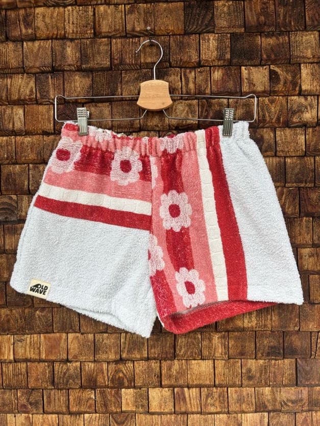 Vorderansicht der Upcycling-Frotteeshorts „Retro Stripes ‘77“ von Old Wave auf einem Kleiderbügel vor einer rustikalen Holzwand. Das asymmetrische Patchwork-Design kombiniert hellgraues oder weißes Frottee mit einem auffälligen, rot-weiß gestreiften Retro-Muster, das mit stilisierten Blütenmotiven verziert ist. Das linke Hosenbein trägt das Old Wave-Logo am Saum.