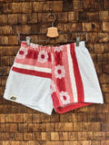 Vorderansicht der Upcycling-Frotteeshorts „Retro Stripes ‘77“ von Old Wave auf einem Kleiderbügel vor einer rustikalen Holzwand. Das asymmetrische Patchwork-Design kombiniert hellgraues oder weißes Frottee mit einem auffälligen, rot-weiß gestreiften Retro-Muster, das mit stilisierten Blütenmotiven verziert ist. Das linke Hosenbein trägt das Old Wave-Logo am Saum.