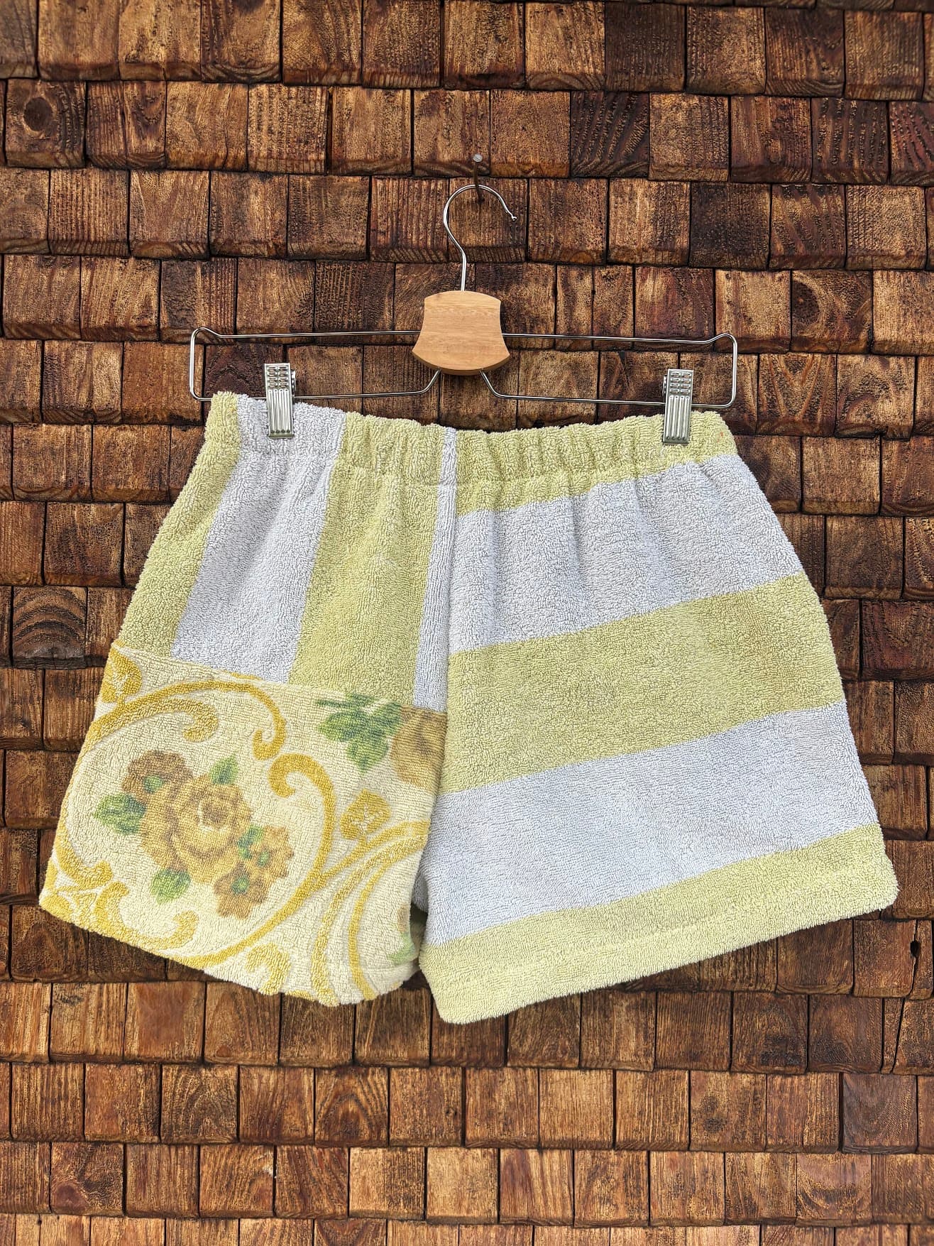 Rückansicht der Upcycling-Frotteeshorts „Sage Brocade ‘78“ von Old Wave auf einem Kleiderbügel vor einer Holzwand. Das asymmetrische Patchwork-Design zeigt auf der rechten Gesäßhälfte breite, horizontale Blockstreifen in Weiß und softem Gelb. Die linke Gesäßhälfte setzt im unteren Bereich das edle, gelb-goldene Vintage-Blumenmuster fort, das im oberen Teil durch vertikale, hellgelbe Stoffsegmente ergänzt wird.