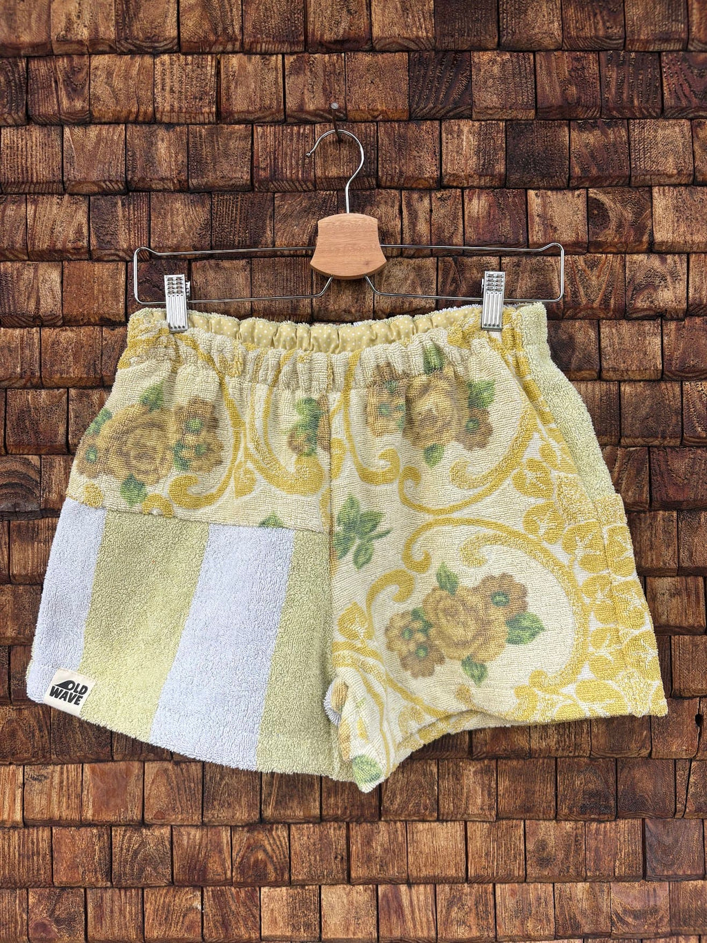 Vorderansicht der Upcycling-Frotteeshorts „Sage Brocade ‘78“ von Old Wave auf einem Kleiderbügel vor einer rustikalen Holzwand. Das elegante Retro-Design kombiniert ein linkes Hosenbein mit einem opulenten, gelb-goldenen Brokat- und Blumenmuster auf hellem Grund mit einem rechten Hosenbein im dezenten Streifen-Look in Pastellgelb und Weiß samt Logo-Patch am Saum.