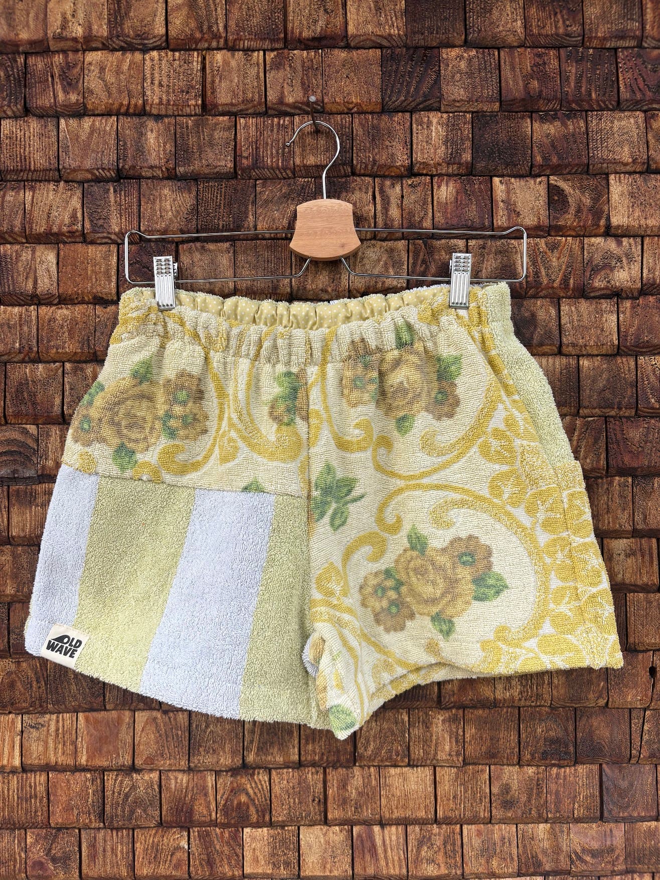 Vorderansicht der Upcycling-Frotteeshorts „Sage Brocade ‘78“ von Old Wave auf einem Kleiderbügel vor einer rustikalen Holzwand. Das elegante Retro-Design kombiniert ein linkes Hosenbein mit einem opulenten, gelb-goldenen Brokat- und Blumenmuster auf hellem Grund mit einem rechten Hosenbein im dezenten Streifen-Look in Pastellgelb und Weiß samt Logo-Patch am Saum.