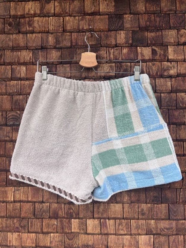 Rückansicht der Upcycling-Frotteeshorts „Salty Grid ‘90“ von Old Wave auf einem Kleiderbügel vor einer Holzwand. Die Rückseite präsentiert ein asymmetrisches Design mit einer schlichten, unifarbenen linken Gesäßhälfte aus weißem Frottee, während die rechte Stoffhälfte mit einem großkarierten Gitter- und Streifenmuster in Hellblau und Salbeigrün gestaltet ist.