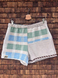 Vorderansicht der Upcycling-Frotteeshorts „Salty Grid ‘90“ von Old Wave auf einem Kleiderbügel vor einer rustikalen Holzwand. Das maritime Patchwork-Design zeigt ein linkes Hosenbein mit breiten, horizontalen Blockstreifen in Hellblau und Salbeigrün auf weißem Grund sowie dem eingenähten Old Wave-Logo am Saum, kombiniert mit einem rechten Hosenbein aus vorwiegend weißem Vintage-Frottee, das am Bund grüne und blaue Streifenakzente besitzt.