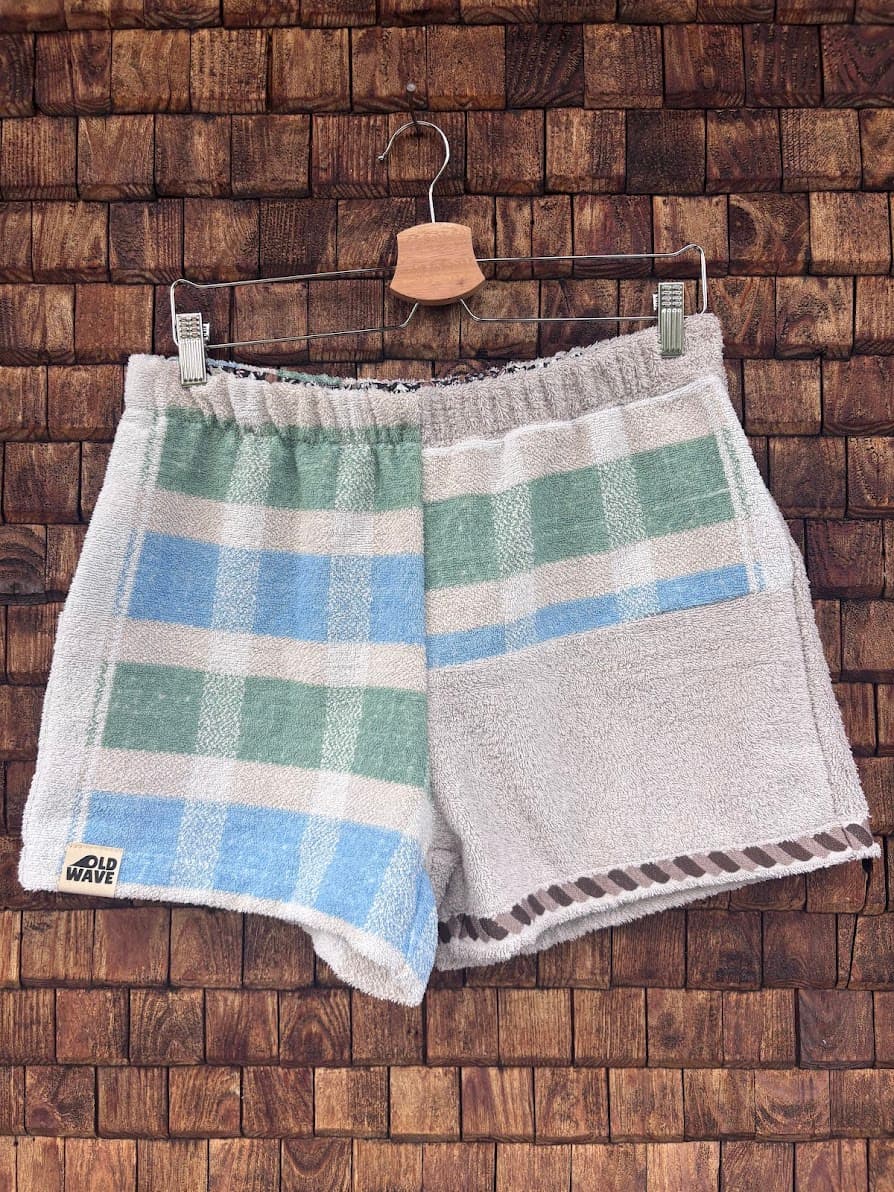 Vorderansicht der Upcycling-Frotteeshorts „Salty Grid ‘90“ von Old Wave auf einem Kleiderbügel vor einer rustikalen Holzwand. Das maritime Patchwork-Design zeigt ein linkes Hosenbein mit breiten, horizontalen Blockstreifen in Hellblau und Salbeigrün auf weißem Grund sowie dem eingenähten Old Wave-Logo am Saum, kombiniert mit einem rechten Hosenbein aus vorwiegend weißem Vintage-Frottee, das am Bund grüne und blaue Streifenakzente besitzt.